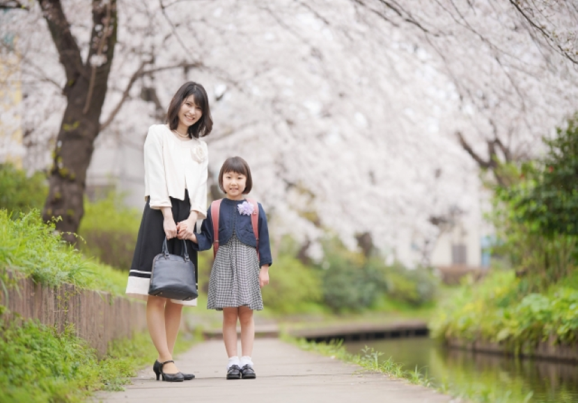 桜並木を歩くスーツの母と入学生